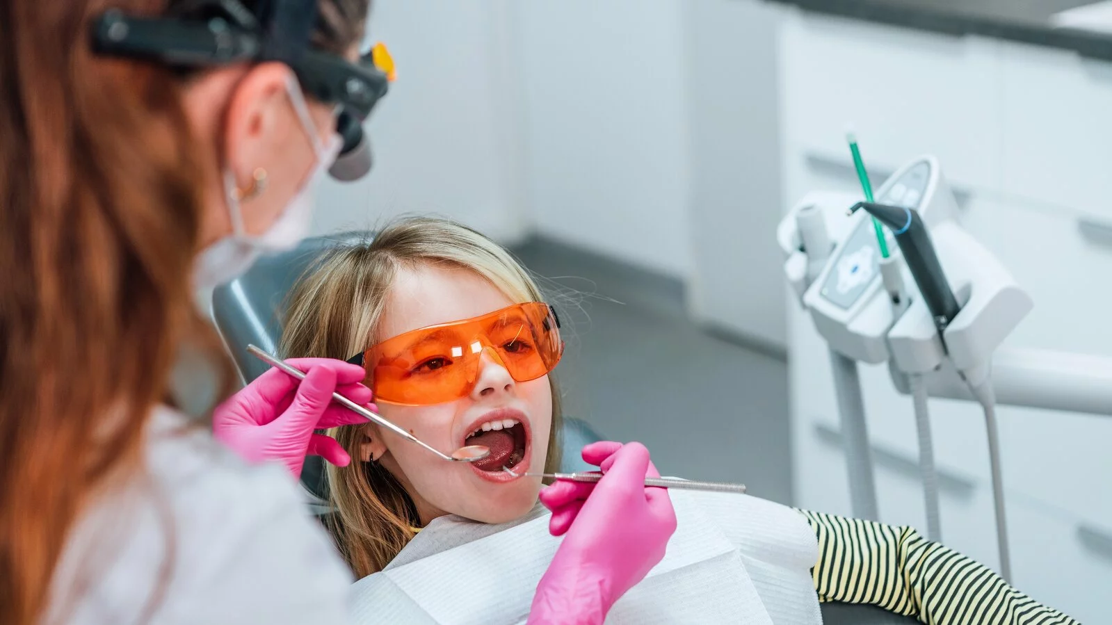 Child in protective glasses sitting in operatory chair while hygienist in pink gloves with excavator and mirror examines.