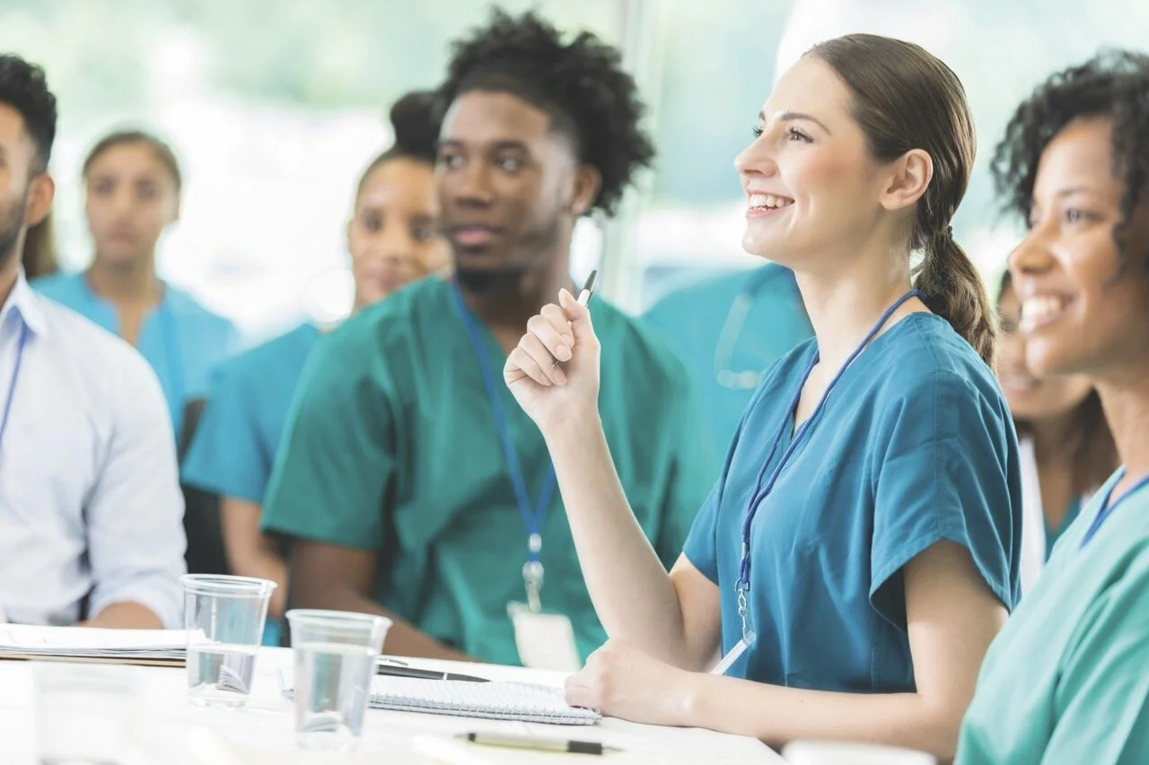 Cheerful young Hispanic female nurse attentively listens to a speaker during a medical seminar. Her colleagues are sitting next to her and are in the background. 2025/02/iStock-858087832-Large.jpeg 